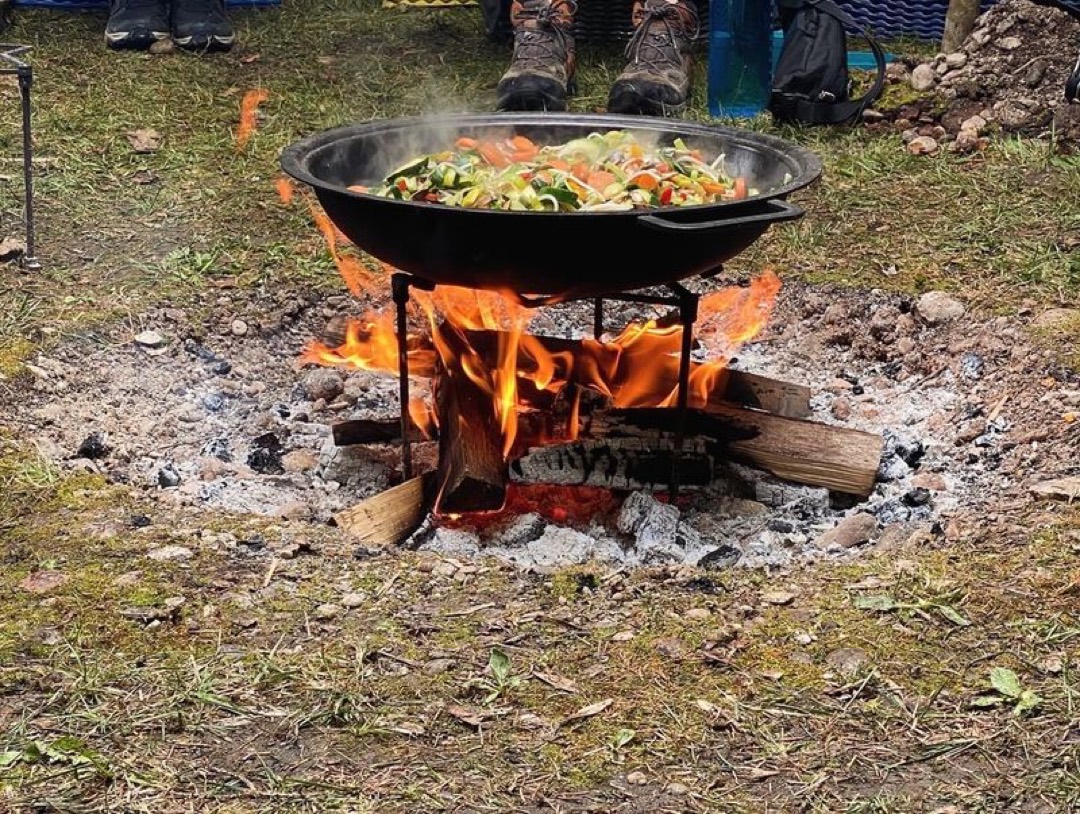 Gemüsewok mit Zuchhetti, Karotten, Zwiebeln in grosser Pfanne über einem Feuer am kochen im Hintrgrund Wanderschuhe