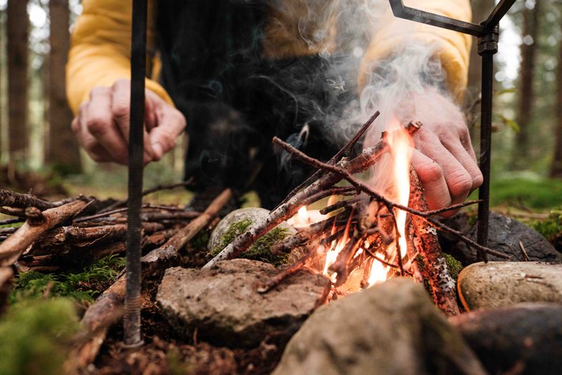 Nahaufnahme von zwei Händen die ein Feuer mit kleinen Ästen mitten im Wald entfachen